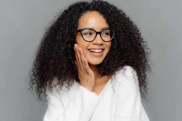 Headshot of beautiful dark skinned female model touches cheek gently, has charming smile on face, looks happily away, has curly Afro hair, wears spectacles and white jumper, isolated on grey wall - Photo, Image