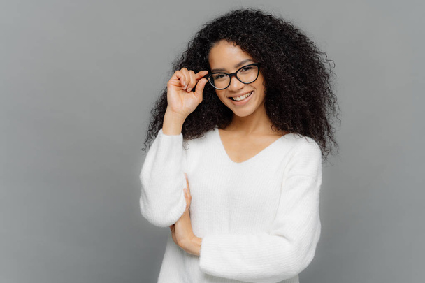 Portrait of happy woman with curly hair, kepes hands on frame of glasses, has gentle smile, wears white jumper, isolated over grey background. Copy space for your advert. Delighted student indoor - Photo, Image