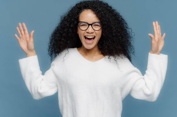 Overjoyed mixed race woman with curly hair, raises hands, exclaims from positive emotions, keeps mouth opened, dressed in white sweater, stands against blue background. Facial expressions concept - Photo, Image