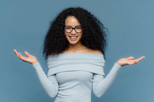 Waist up shot of happy young crisp Afro American woman raises both hands, pretends holding something, wears blue sweater, has naked shoulders, optical glasses, isolated over blue background. - Photo, Image