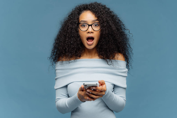 Удивлённая темнокожая женщина с афро-стрижкой, пользуется мобильным телефоном, читает пост в интернете, шокирована последними новостями, носит очки и свитер, изолирован на синем фоне, получает обратную связь
. - Фото, изображение