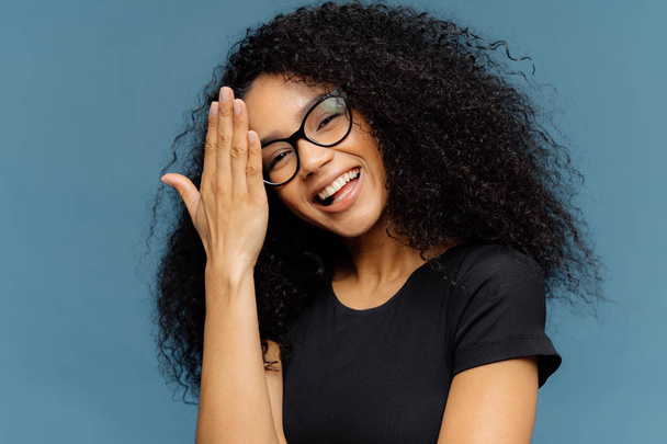 Happy Afro American woman touches forehead, tilts head, smiles happily at camera, has fun indoor, wears optical spectacles and casual black t shirt, isolated over blue background. Emotions concept - Photo, Image