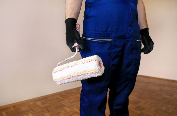 Construction worker at construction site with protective uniform, construction service tool, equipment and instrument. He in blue coverall and holds paint roller brush for wall painting in hand. - Photo, Image