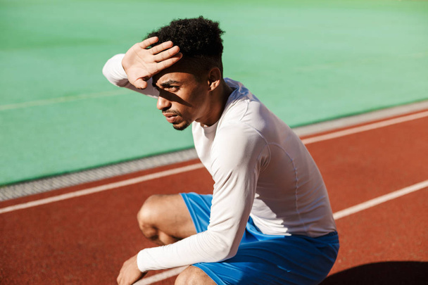Молодой вдумчивый афроамериканский спортсмен сидит на своих ягодицах, закрывая лицо от солнца на городском стадионе.
 - Фото, изображение