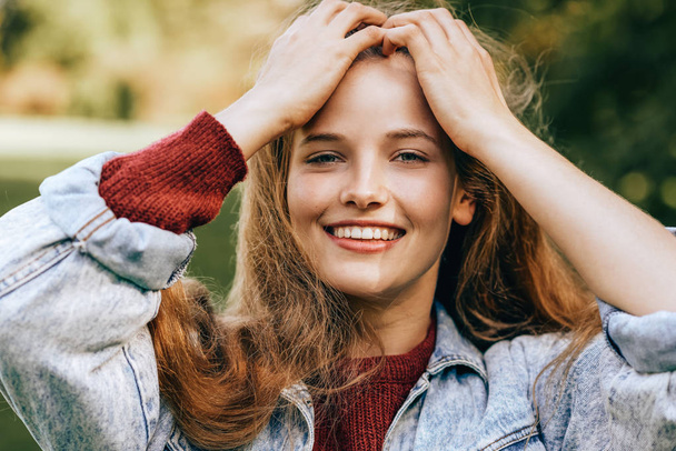 Close-up portrait of beautiful happy young woman smiling broadly with healthy toothy smile, wearing red sweater and denim jacket, posing on nature background in the park. People, travel, lifestyle - Foto, imagen