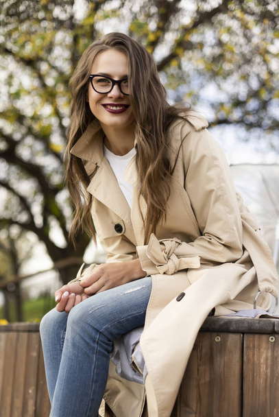 Beautiful smiling girl wearing a light cloak, jeans and a glasse - Foto, imagen