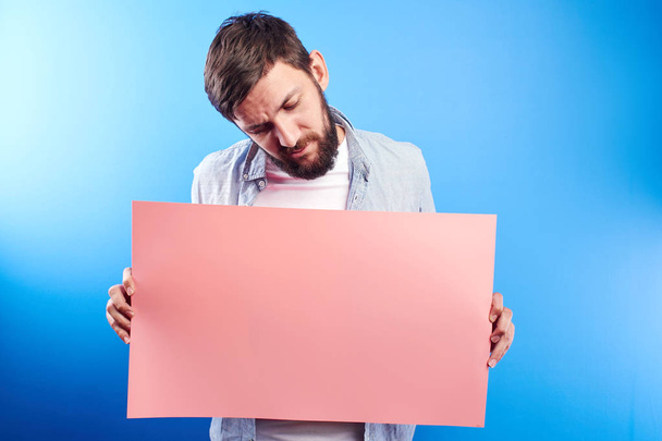 Handsome bearded caucasian man holding blank pink poster board in hands with copy space for text or advertising inscription isolated in blue studio - Foto, Bild