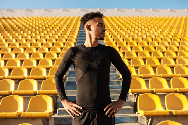 Jovem e atraente esportista afro-americano confiantemente olhando de lado entre os assentos vazios do estádio
  - Foto, Imagem