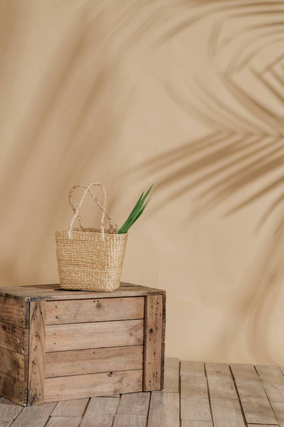 Vertical shot of a straw bag, with a green plant inside, on a wooden box, with a cream background - Photo, image