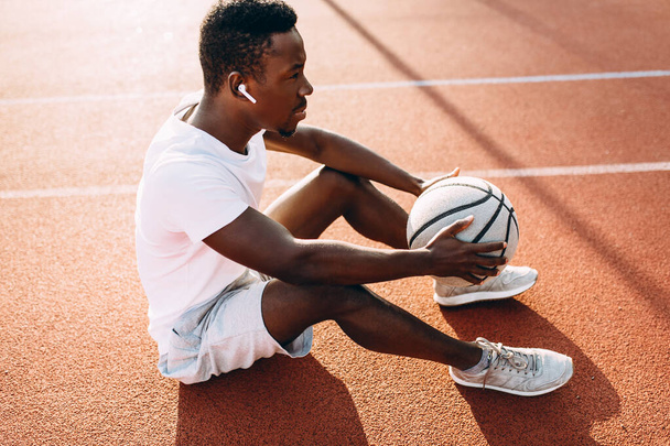 Sports a tired African-American sports person is resting on the basketball court with the ball - Фото, зображення