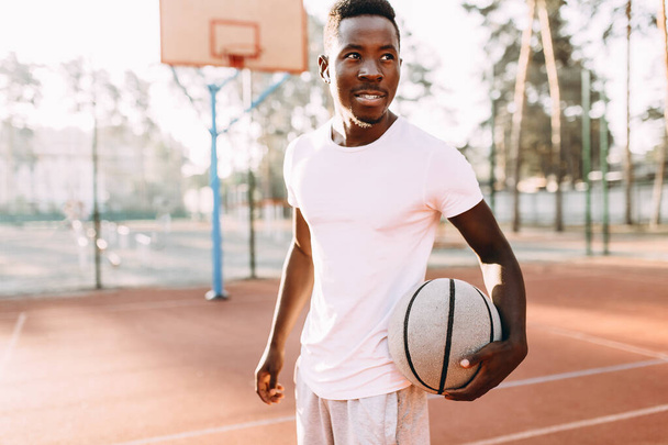 Desportivo jovem afro-americano de pé na quadra ao amanhecer com uma bola de basquete
 - Foto, Imagem