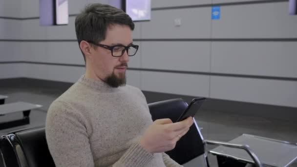 man in glasses types on smartphone in waiting room - Felvétel, videó