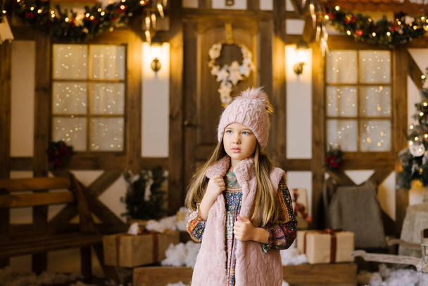 Viiden tai kuuden vuoden tyttö vaaleanpunaisessa neulotussa hatussa, jossa on pompom vaaleanpunaisessa turkisliivissä, seisoo joulutalon taustalla.
 - Valokuva, kuva