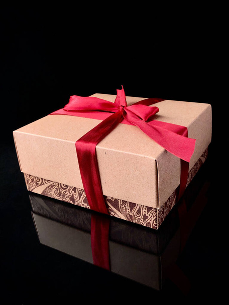 Gift in a box with a red ribbon on a black background. Beige gift box with a red satin ribbon and bow. - Photo, Image