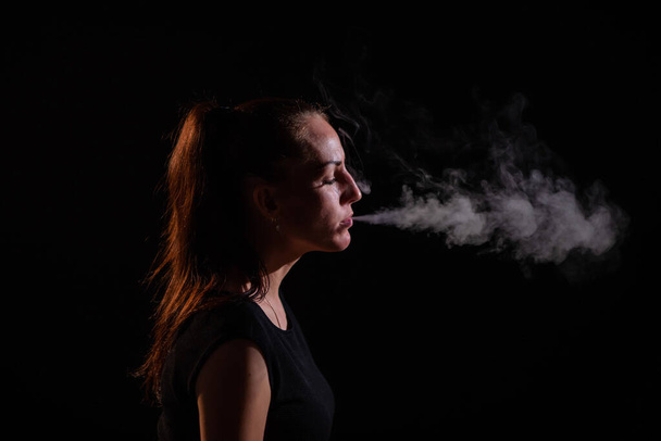 The brunette smokes an electronic cigarette on a black background, releases thick smoke from her mouth. Portrait of a woman smoking a vape, hovering. - Фото, изображение