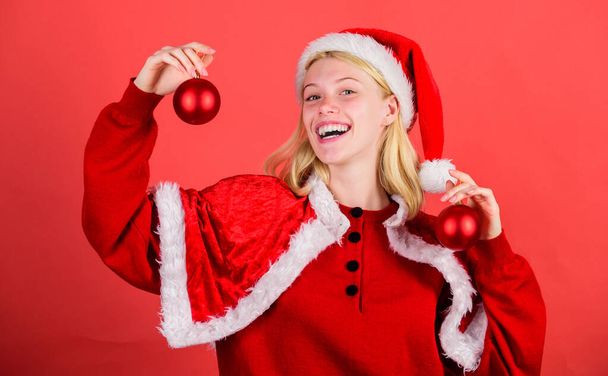 Christmas preparation concept. Favorite time year christmas. Christmas fun. Enjoy celebration with costume and decor. Girl happy wear santa costume celebrate christmas hold ball decor red background - Photo, image