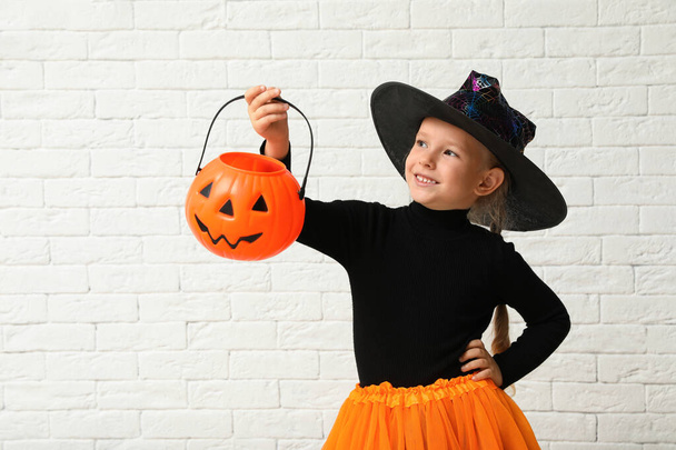 Cute little girl with pumpkin candy bucket wearing Halloween costume near white brick wall - Fotó, kép