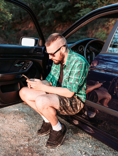 Cute bearded man with sunglasses sitting at car door looking at his phone. - Zdjęcie, obraz