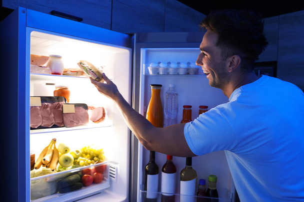 Man taking sandwich out of refrigerator in kitchen at night - Фото, зображення
