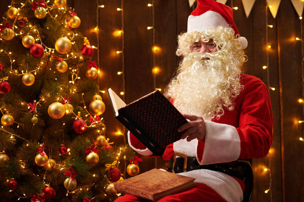Libro di lettura di Babbo Natale, seduto al coperto vicino all'albero di Natale decorato con luci Buon Natale e Buone Feste
! - Foto, immagini