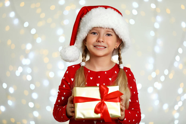 Happy little child in Santa hat with gift box against blurred festive lights. Christmas celebration - Photo, image