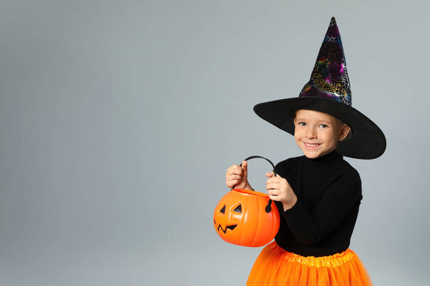 Cute little girl with pumpkin candy bucket wearing Halloween costume on grey background, space for text - 写真・画像