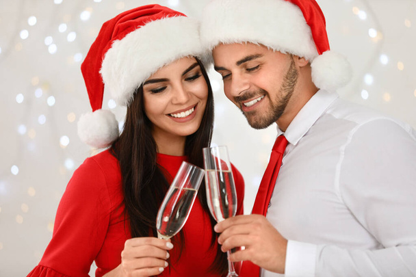 Happy young couple in Santa hats with glasses of champagne against blurred festive lights. Christmas celebration - Foto, imagen