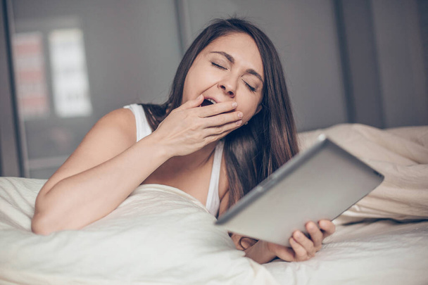 Young woman with tablet in her hands yawning in bed - Photo, image