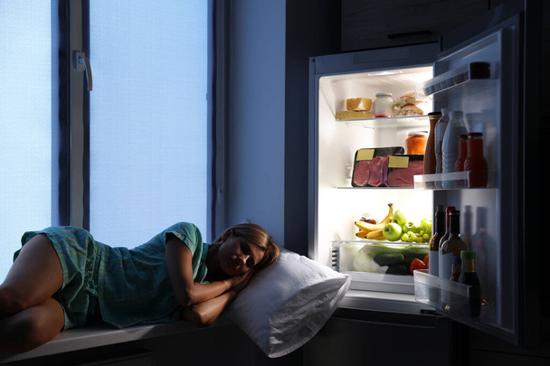 Woman sleeping on window sill near open refrigerator in kitchen at night - Fotografie, Obrázek