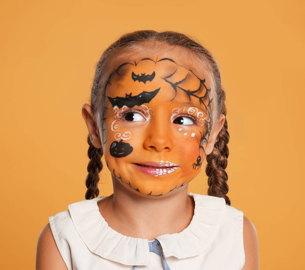 Cute little girl with face painting on orange background - Фото, изображение