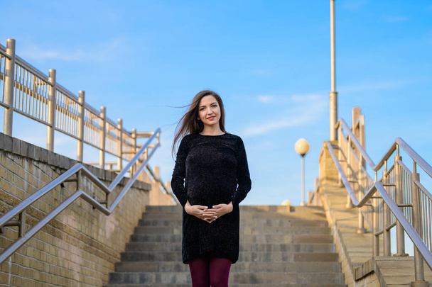 έγκυος γυναίκα στον γαλάζιο ουρανό. Πορτρέτο μιας νεαρής εγκύου ενάντια σε έναν καταγάλανο ουρανό, αστική εγκυμοσύνη, όμορφη και ευτυχισμένη έγκυος μελαχρινή με μακριά μαλλιά - Φωτογραφία, εικόνα