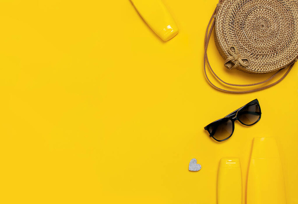 Female summer fashion background. Clothes and accessories on yellow background. Blue hat, denim shorts, round rattan bag, sandals, sunglasses, sunscreen, phone. Flat lay top view fashion blog concept - Photo, Image