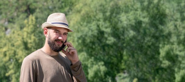 A bearded guy in a hat speaks on the phone and looks sadly into the frame. The lack of telephone communications in nature. The concept of tourism, technical problem. Copy space. Template for a banner. - Foto, imagen