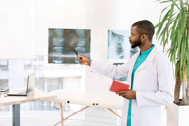 Young male african american health care worker doctor in white coat standing in modern clinic office and looking at patient x-ray - Fotoğraf, Görsel