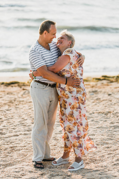 Romantic adult couple standing together on the beach. Mature pair relax at the seaside on holiday. Happy familiy walking and holding hands near sea. Husband embracing wife. Love story near the sea - Foto, imagen