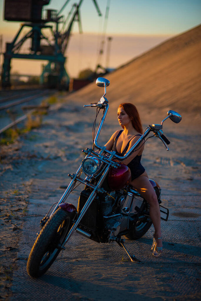 Red-haired woman in sexy lingerie in high heels sits on a motorcycle. Attractive red-haired girl sits on a motorcycle at sunset - Photo, image