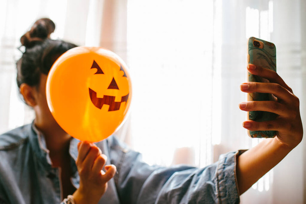 Witch holding a pumpkin balloon with her hand, covering her face and taking a photo. Young woman celebrating halloween. Helium party balloons. - Фото, зображення