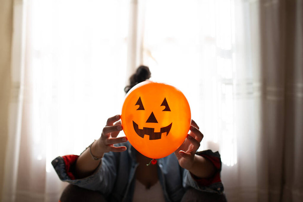 Bruja sosteniendo un globo de calabaza con sus manos y cubriendo su cara. Mujer joven celebrando halloween. Globos de fiesta de helio
. - Foto, Imagen