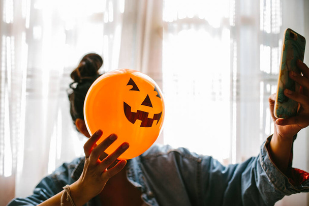 Witch holding a pumpkin balloon with her hand, covering her face and taking a photo with mobile phone. Young woman celebrating halloween. Helium party balloons. - Φωτογραφία, εικόνα