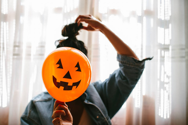 Witch holding a pumpkin balloon with her hand and covering her face. Young woman celebrating halloween. Helium party balloons. - 写真・画像