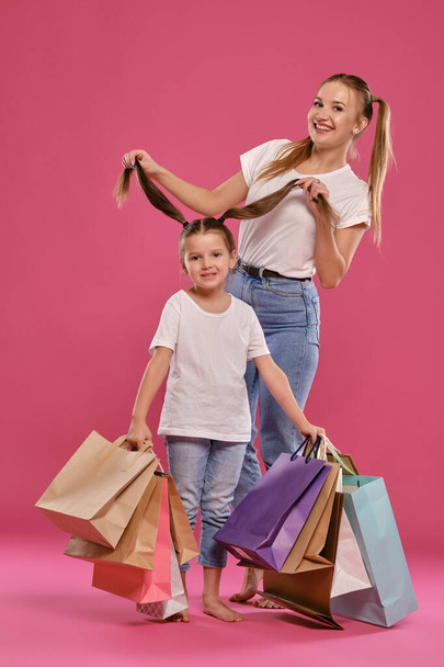 Mom and daughter with ponytails, dressed in white t-shirts and blue jeans are posing against pink background with packages in hands. Full length. - Photo, image