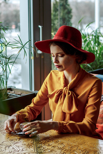 girl in a cafe with a cup of coffee and a hat.portrait of sensual young girl wearing floppy hat and blouse with bow. Beautiful brunette woman in cafe holding cup of coffee - Фото, изображение