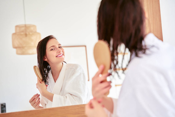 Photo of brunette combing her hair in front of bathtub mirror - Fotoğraf, Görsel