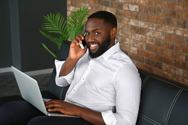 Komea afroamerikkalainen liikemies kannettavan tietokoneen kanssa puhuu matkapuhelimella toimistossa
 - Valokuva, kuva