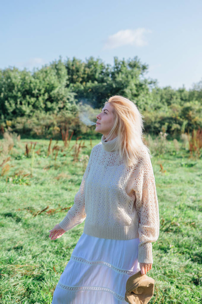 Beautiful blond woman staying and smocking cigarette in a green field - Foto, afbeelding