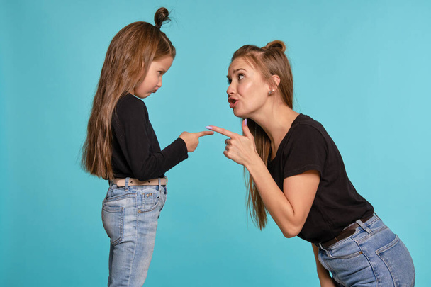 Mom and daughter with a funny hairstyles, dressed in black shirts and blue denim jeans are posing against a blue studio background. Close-up shot. - Foto, imagen