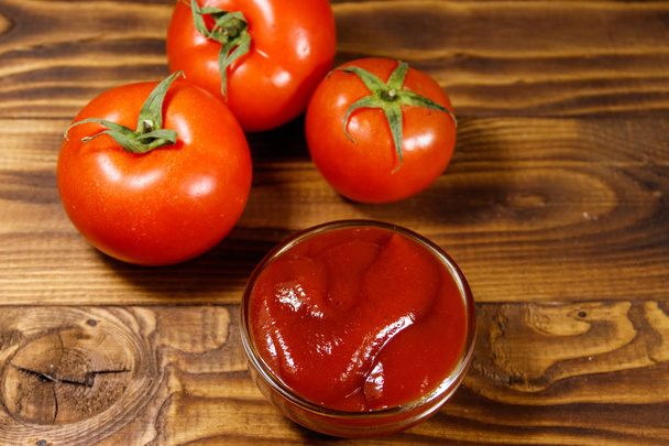 Lasikulho ketsuppia tai tomaattikastiketta ja tuoreita kypsiä tomaatteja puupöydällä
 - Valokuva, kuva