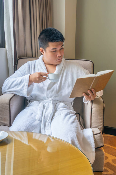 Asian businessman wearing bathrobe and reading book at hotel room - Фото, изображение