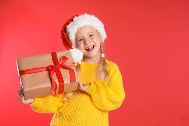 Happy little child in Santa hat with gift box on red background. Christmas celebration - Zdjęcie, obraz