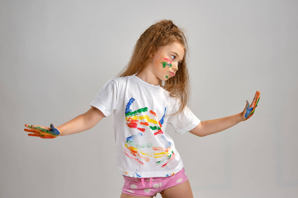 Menina em branco pintado t-shirt está posando de pé isolado em branco e gesticulating com ela colorido em diferentes tintas palmas e rosto. Estúdio de arte. Close-up. - Foto, Imagem